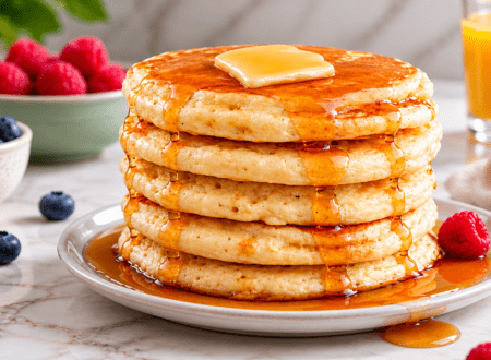 Stack of fluffy vegan pancakes topped with creamy glaze and pecans on a white plate against a dark cozy background.