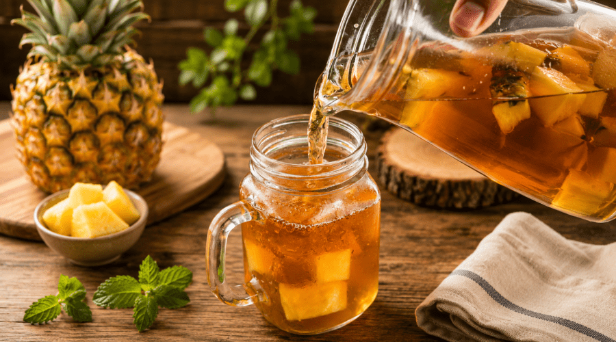 Glass of iced pineapple tea garnished with pineapple wedges, lemon slices, mint leaves, and ice cubes.